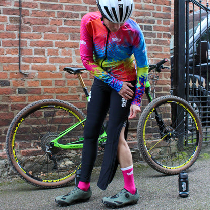 Woman wearing pink and blue multicoloured cycling jersey zipping up a pair of black Zip-off tights, standing in front of a MTB  bike against a brick wall.